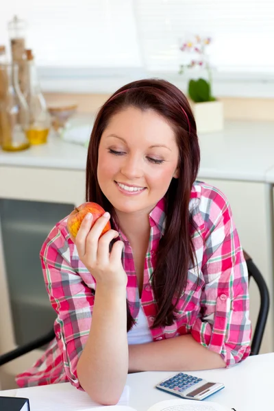 Mujer caucásica sonriente sosteniendo una manzana sentada en la cocina —  Fotos de Stock