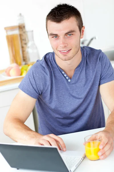 Чарівний молодий чоловік, використовуючи свій ноутбук, тримає апельсиновий сік в — стокове фото