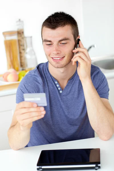 Attractive young man talking on phone holding a card in the kitc — Stock Photo, Image