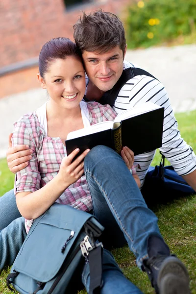 Smiling couple of students reading a book sitting on grass — Stock Photo, Image