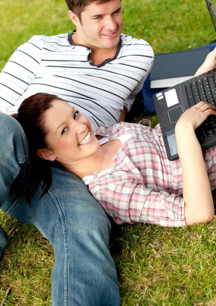 Couple of cheerful students using a laptop lying on the grass — Stock Photo, Image