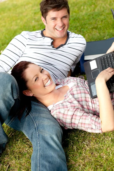 Couple of happy students using a laptop lying on the grass — Stock Photo, Image