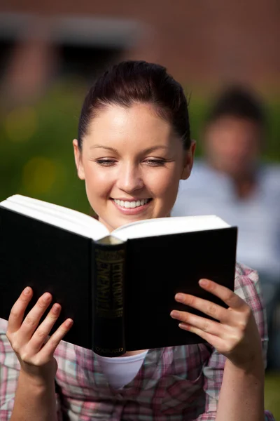 Χαμογελώντας φοιτήτριας, διαβάζοντας ένα βιβλίο που κάθεται στο γρασίδι στην — Φωτογραφία Αρχείου