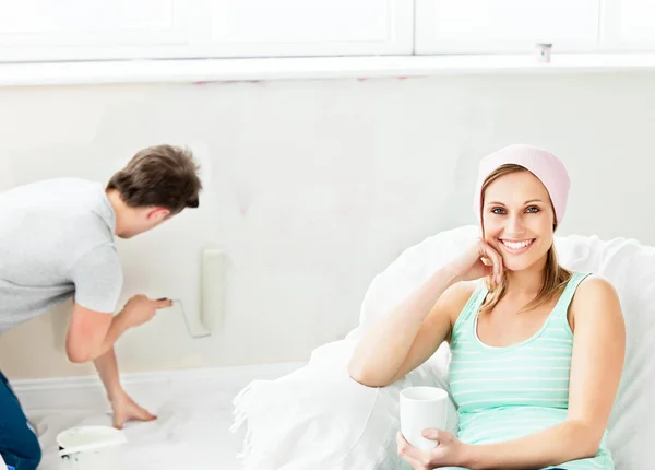 Smiling caucasian woman relaxing on a sofa while boyfriend paint — Stock Photo, Image