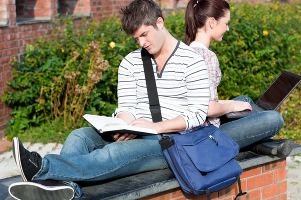 Un par de estudiantes usando un portátil y leyendo un libro sentado —  Fotos de Stock