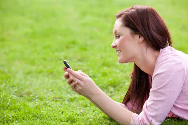 Blij jonge vrouw schrijven van een bericht op haar telefoon zittend op het gras — Stockfoto