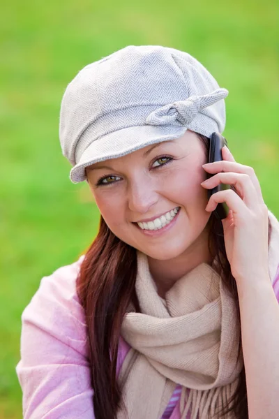Pretty young woman wearing cap and scarf talking on phone on the grass — Stock Photo, Image