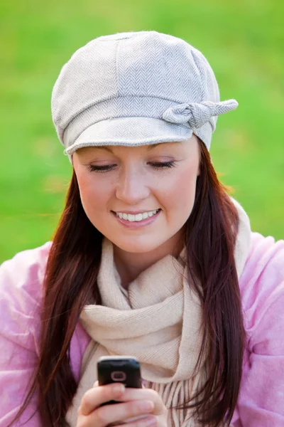 Jolie jeune femme portant casquette et écharpe envoyant un message avec son téléphone portable sur l'herbe — Photo