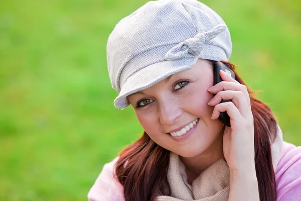 Smiling young woman wearing cap and scarf talking on phone on the grass — Stock Photo, Image