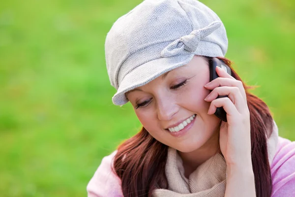 Smiling young woman wearing cap and scarf talking on phone on the grass — Stock Photo, Image