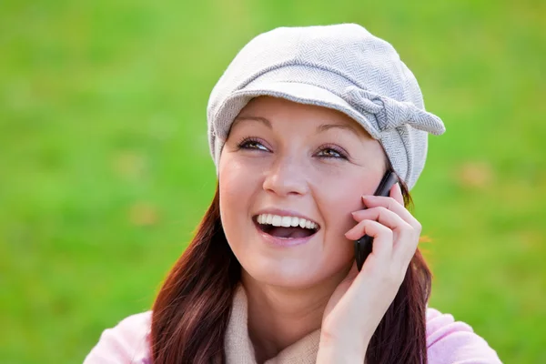 Смеющаяся молодая женщина в шапке и шарфе разговаривает по телефону на траве — стоковое фото