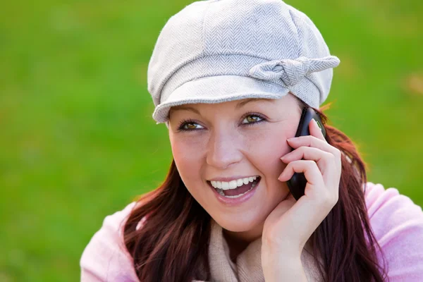 笑着年轻女子戴帽和围巾在草地上，电话上谈了 — 图库照片