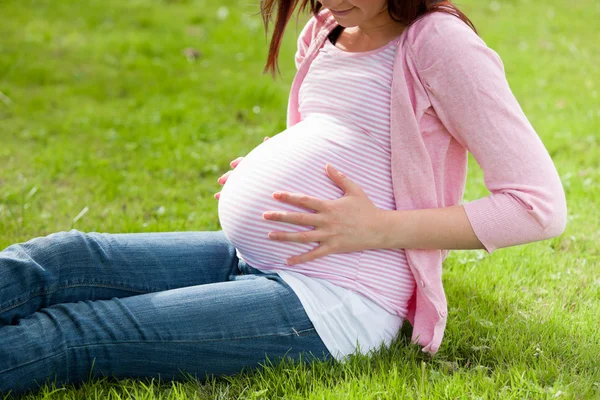 Беременная женщина держит живот, сидя на траве. — стоковое фото