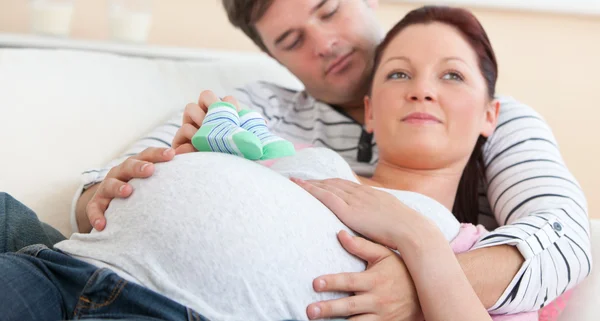 Söt framtida mamma liggande med make i soffan håller baby sko — Stockfoto
