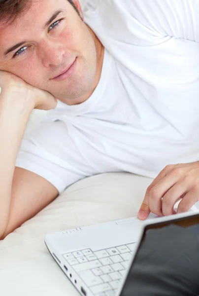 Bonito homem escrevendo em seu laptop deitado em seu quarto — Fotografia de Stock