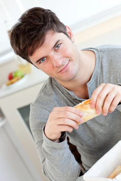 Ελκυστικό νεαρό άνδρα που τρώει ψωμί, κάθεται στην κουζίνα του — Φωτογραφία Αρχείου