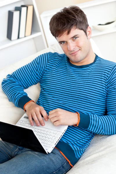 活泼开朗的年轻人坐在沙发上躺在他的笔记本上工作 — 图库照片