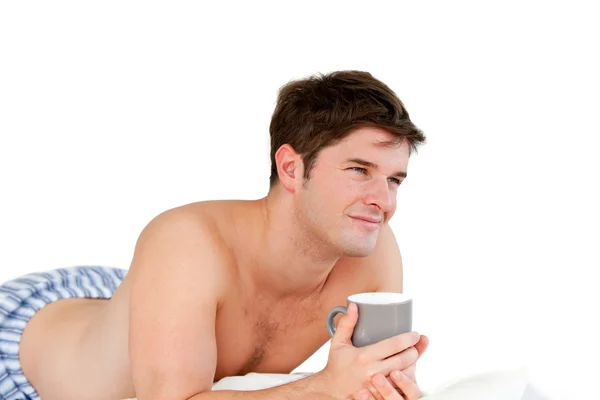 Uomo sorridente che tiene una tazza di caffè sdraiato sul suo letto durante il — Foto Stock