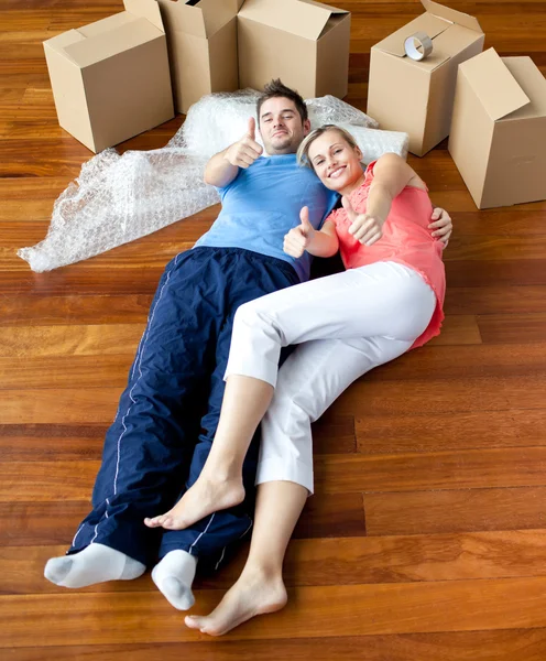 Счастливая пара лежит на полу в своем новом доме и делает большие пальцы - — стоковое фото
