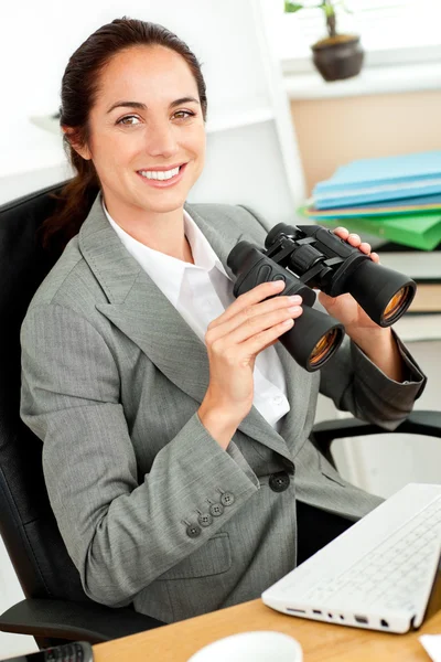Smiling hispanic businesswoman holding binoculars sitting at her — Stock Photo, Image