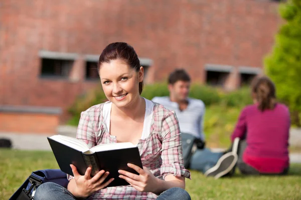 Delighted female student reading a book sitting on grass — Stock Photo, Image