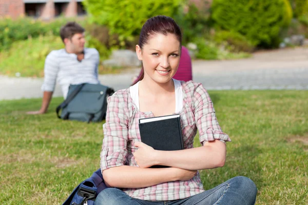 Sorrindo estudante do sexo feminino lendo um livro sentado na grama — Fotografia de Stock