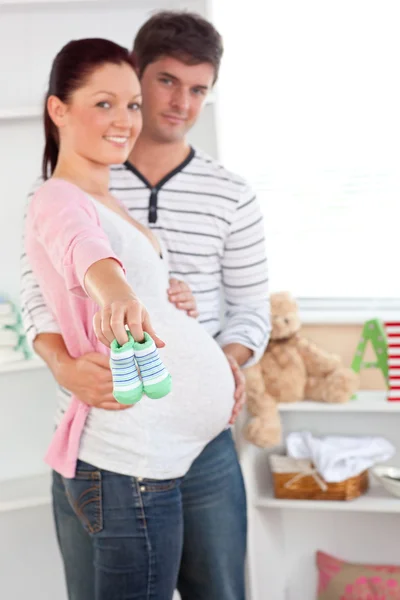 Mujer embarazada alegre sosteniendo zapatos de bebé mientras su marido toca — Foto de Stock