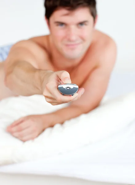 Bonito homem segurando um controle remoto deitado em sua cama — Fotografia de Stock