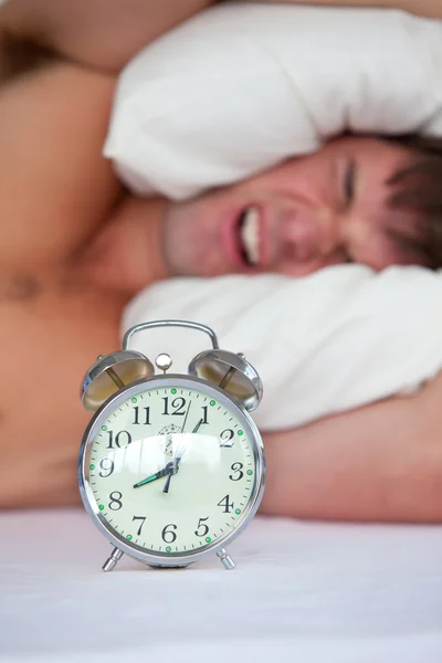 Разъяренный молодой человек лежит в постели и раздражен своим будильником в футболке — стоковое фото