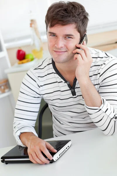 Hombre atractivo hablando por teléfono usando su portátil en la cocina —  Fotos de Stock