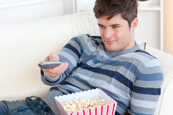 Lumineux jeune homme mangeant du pop-corn et tenant une télécommande couchée sur le th — Photo