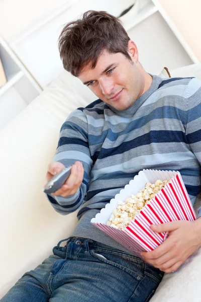 Delighted young man eating popcorn and holding a remote lying on — Stock Photo, Image