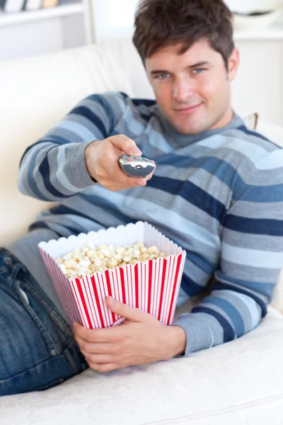 Entspannter junger Mann isst Popcorn und hält eine Fernbedienung auf dem Boden — Stockfoto