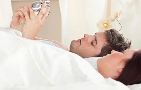 Young caucasian couple holding an alarm clock in bed before slee — Stock Photo, Image