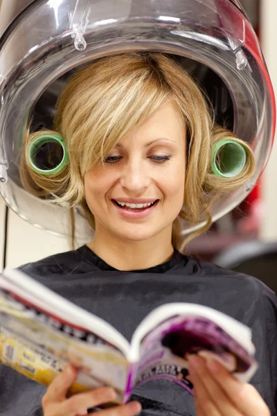 Femme heureuse lisant un magazine avec des bigoudis sous un sèche-cheveux — Photo