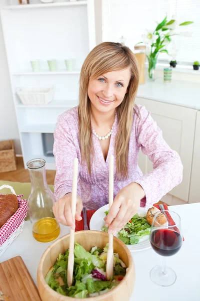 Retrato de una linda mujer sirviendo ensalada sentada en la cocina — Foto de Stock