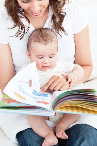 Ευτυχισμένη μητέρα δείχνει ένα βιβλίο για να το μωρό κάθεται στον καναπέ στο h — Φωτογραφία Αρχείου