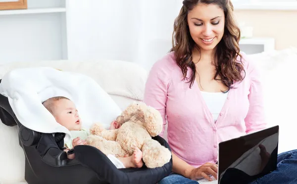 Madre brillante trabajando en un portátil con su bebé sentado junto a — Foto de Stock