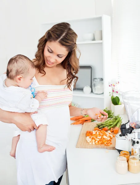 Młoda matka trzyma swojego dziecka podczas przygotowywania marchew na obiad — Zdjęcie stockowe