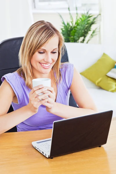 Ελκυστική γυναίκα, κρατώντας ένα φλιτζάνι καφέ και χρησιμοποιώντας φορητό υπολογιστή sittin — Φωτογραφία Αρχείου