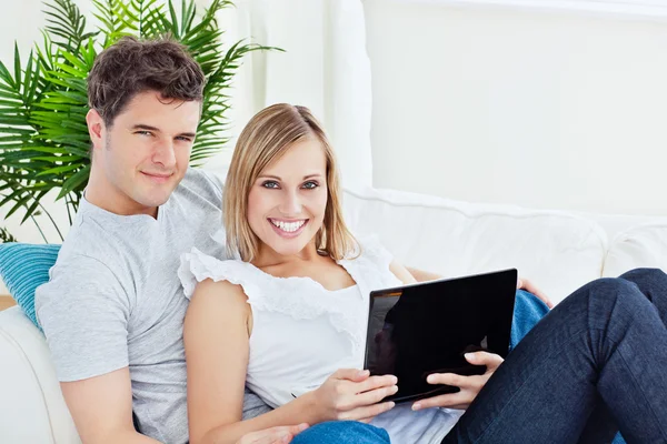 Joven feliz pareja acostada en el sofá con el ordenador portátil sonriendo a la —  Fotos de Stock