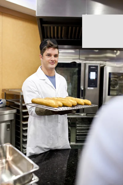 Jovem padeiro segurando baguetes e pães de pé na cozinha — Fotografia de Stock