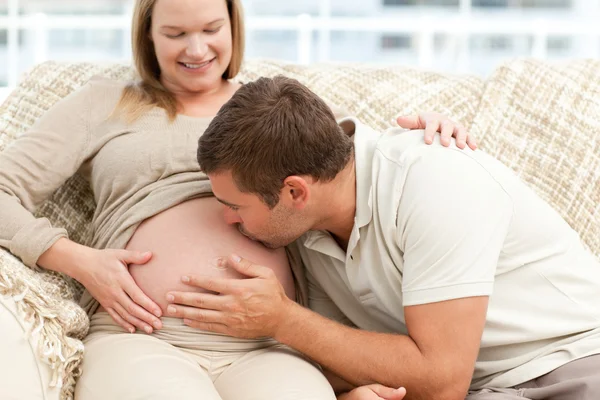 Budoucí tatínek líbat bříško jeho žena sedí na pohovce — Stock fotografie