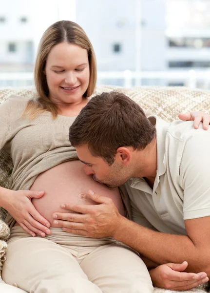 Pozorný člověk líbat jeho manželky břicho při relaxaci na sof — Stock fotografie