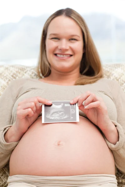 Χαρούμενα έγκυος γυναίκα, κρατώντας ένα υπερηχογράφημα κάθεται στο το sof — Φωτογραφία Αρχείου