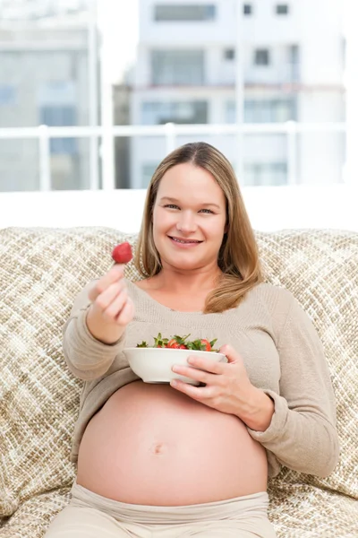 Futur mom with a bowl of strawberries sitting on the sofa — Stockfoto