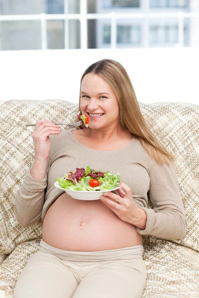 Ευτυχισμένη έγκυος γυναίκα, απολαμβάνοντας μια σαλάτα που κάθεται στον καναπέ — Φωτογραφία Αρχείου