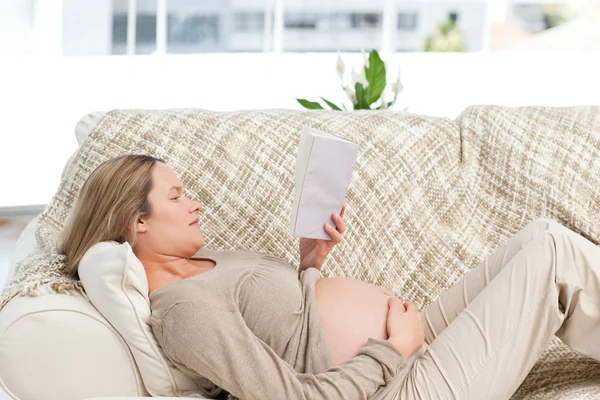 Концентрированная беременная женщина читает книгу, лежащую на диване — стоковое фото