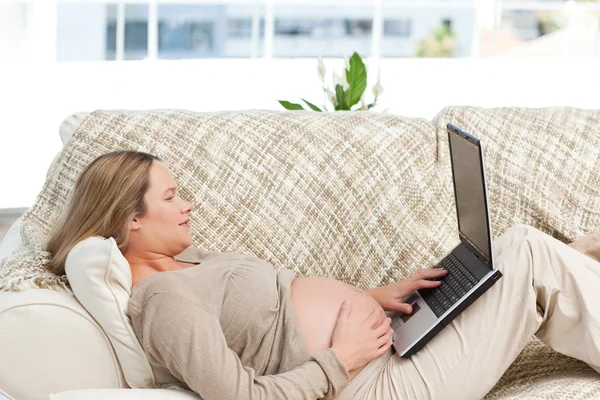 Улыбающаяся беременная женщина с помощью ноутбука лежит на диване — стоковое фото