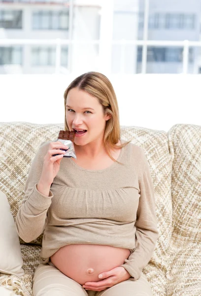 Ziemlich zukünftige Mutter essen einen Schokoriegel sitzt auf dem Sofa — Stockfoto